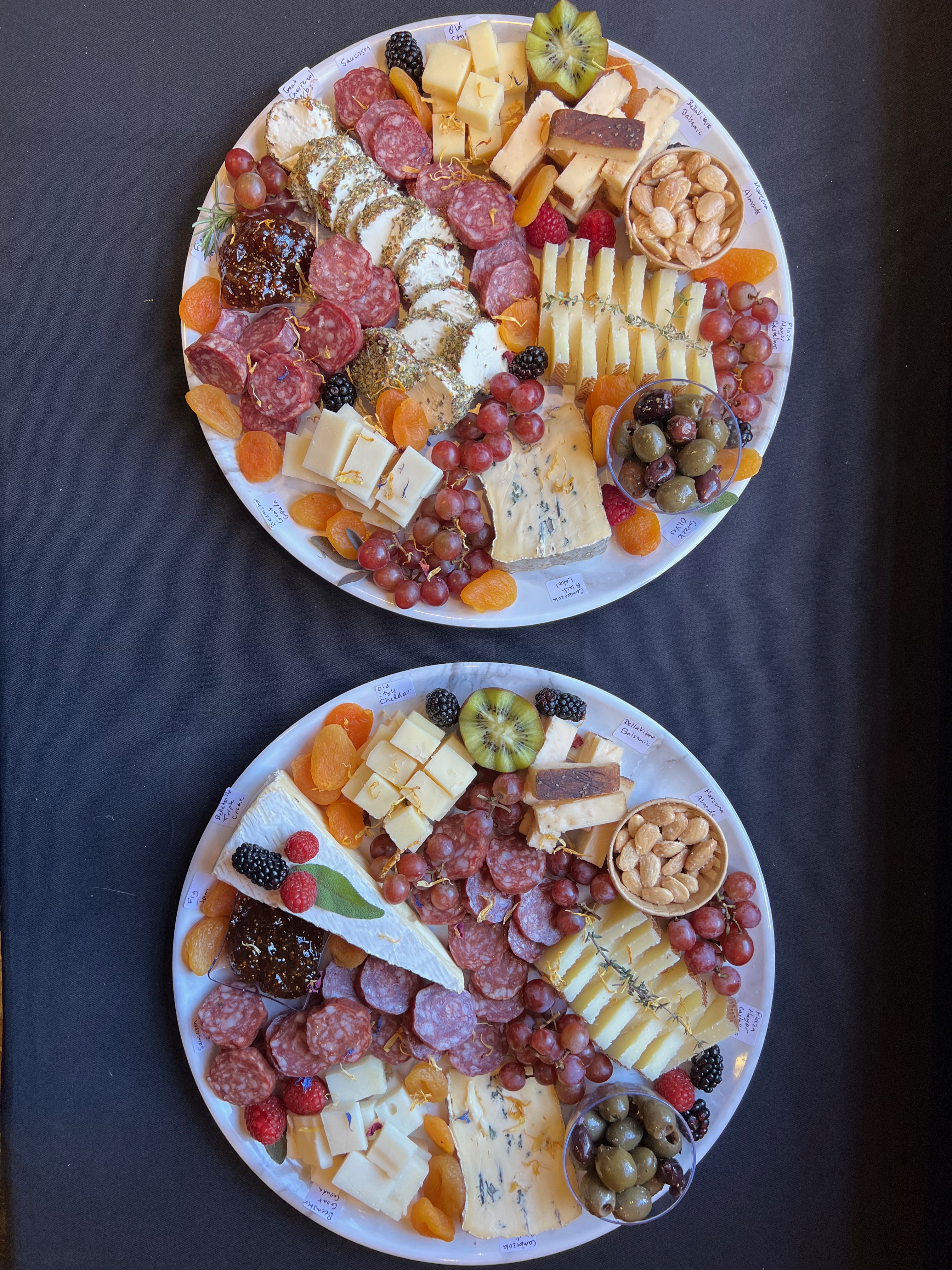 Top-down view of two cheese platters containing an assortment of cheeses, salami, and fruits.